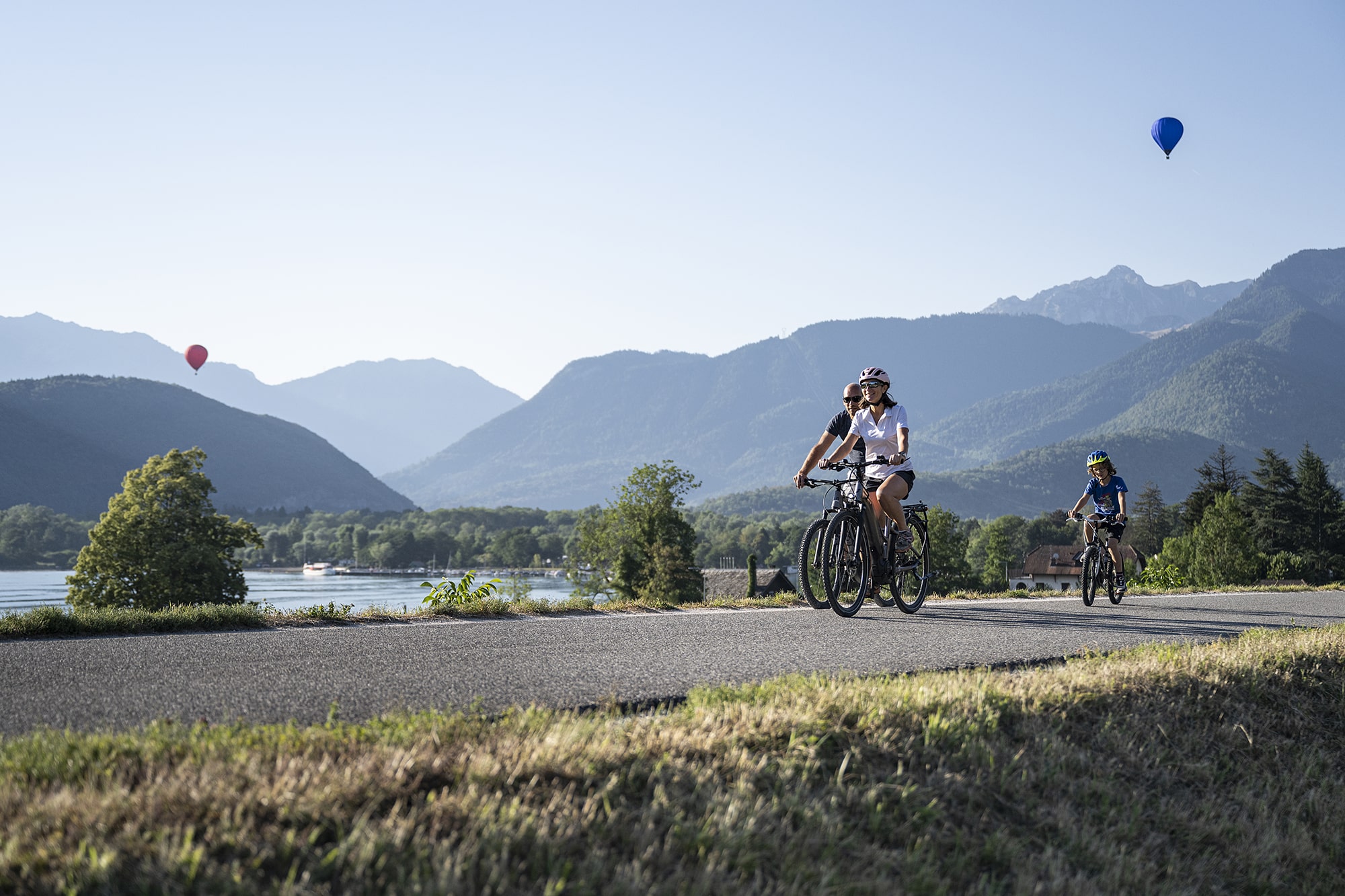 Deux adultes et un enfant à vélo sur la piste cyclable vue lac d'Annecy et montgolfières